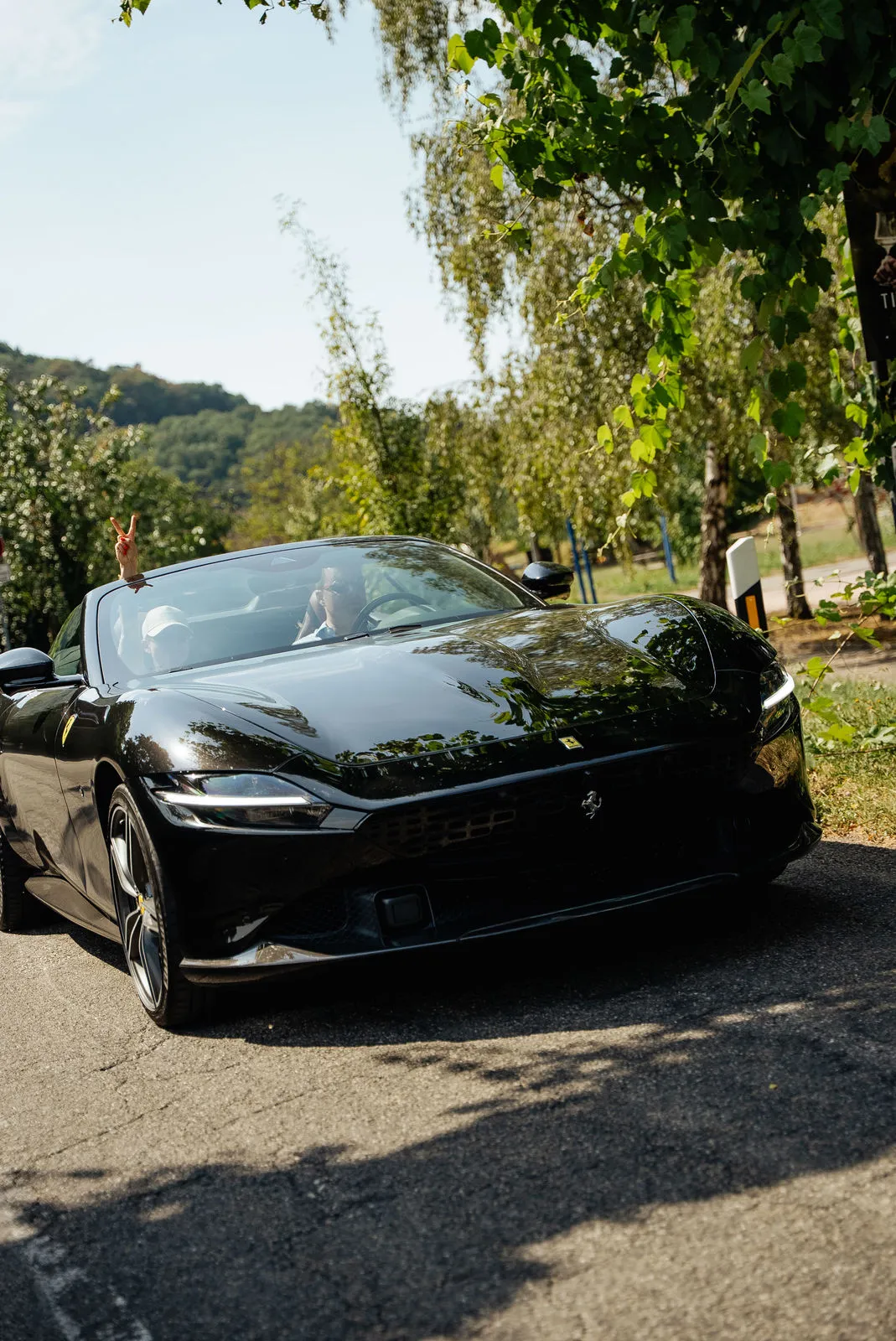 Schwarzer Ferrari Roma Spider auf einer Sportwagenausfahrt, aus dem Fahrzeug winkt eine Hand mit Peace-Zeichen